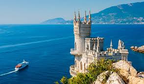 На изображении показан замок Ласточкино гнездо, расположенный на скале над Черным морем в Крыму. Замок имеет готический стиль с башнями и шпилями. На переднем плане видны скалы и зелень, а на заднем плане - море и горы. В море плывет белый катер.